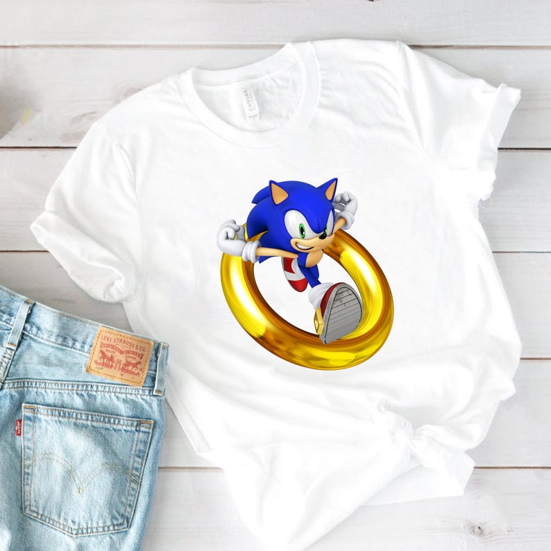 May include: White t-shirt featuring a graphic of Sonic the Hedgehog in a running pose, passing through a golden ring. The character is blue with red shoes. The shirt is paired with light blue denim jeans.