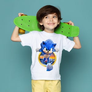 May include: A white t-shirt featuring a blue Sonic the Hedgehog graphic inside a gold circle. The child is holding a green skateboard. The background is a solid teal color.