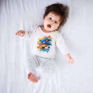 May include: A baby wearing a white long-sleeved onesie with a colorful graphic of Sonic the Hedgehog wearing sunglasses. The baby is also wearing light gray pants with a floral pattern. The baby is lying on a white sheet.