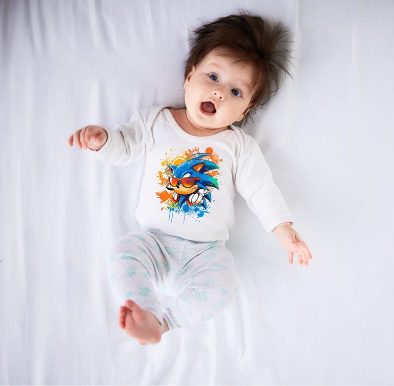 May include: A baby wearing a white long-sleeved onesie with a colorful graphic of Sonic the Hedgehog wearing sunglasses. The baby is also wearing light gray pants with a floral pattern. The baby is lying on a white sheet.