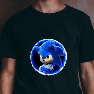 May include: Black t-shirt featuring a blue cartoon character, Sonic the Hedgehog, within a blue glowing circle. The character has blue fur, green eyes, and crossed arms. The shirt is a solid color.