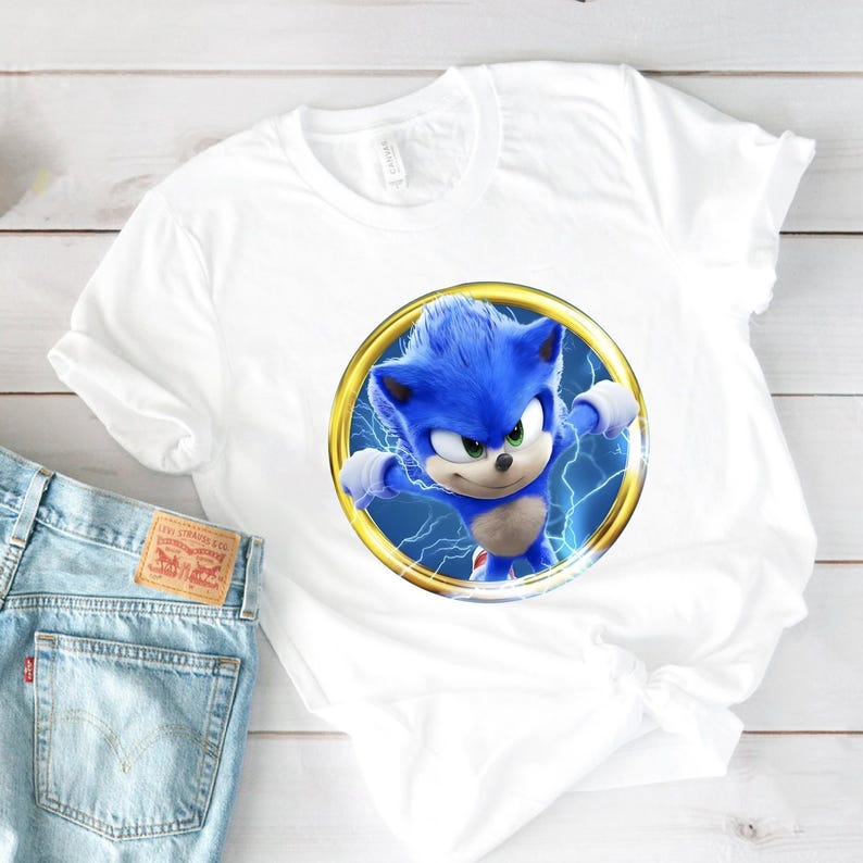 May include: White t-shirt featuring a vibrant graphic of Sonic the Hedgehog in a gold ring with blue lightning. The blue character has a determined expression. The shirt is paired with light blue denim jeans.
