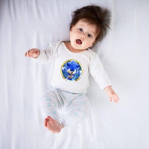 May include: A baby wearing a white long-sleeved onesie with a circular graphic of Sonic the Hedgehog. The graphic features the blue character with yellow lightning bolts. The baby is also wearing light gray and white patterned pants.