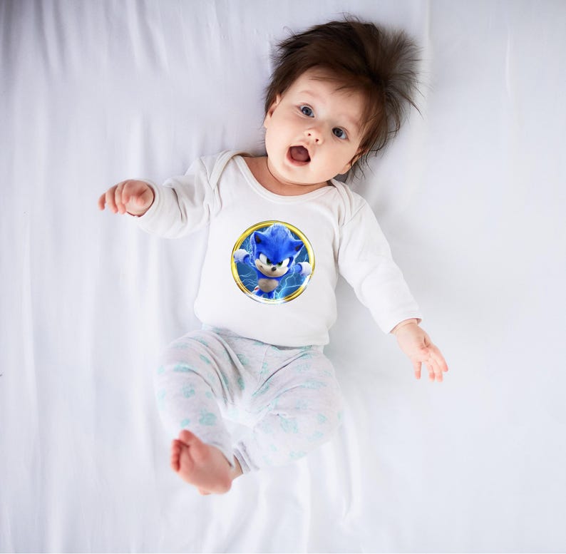 May include: A baby wearing a white long-sleeved onesie with a circular graphic of Sonic the Hedgehog. The graphic features the blue character with yellow lightning bolts. The baby is also wearing light gray and white patterned pants.