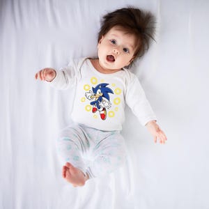 May include: A baby wearing a white long-sleeved onesie with a graphic of Sonic the Hedgehog. The blue character is surrounded by yellow rings. The baby is also wearing light gray and white patterned pants.