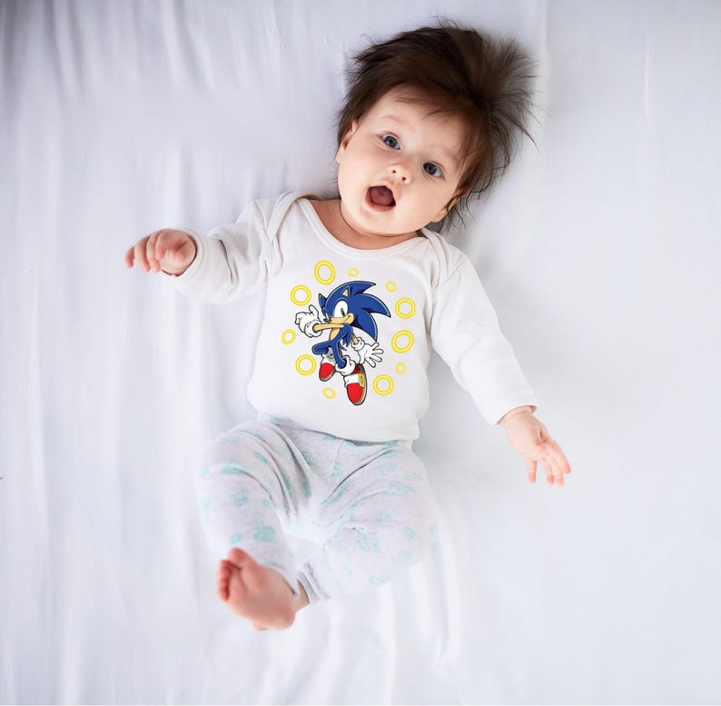 May include: A baby wearing a white long-sleeved onesie with a graphic of Sonic the Hedgehog. The blue character is surrounded by yellow rings. The baby is also wearing light gray and white patterned pants.