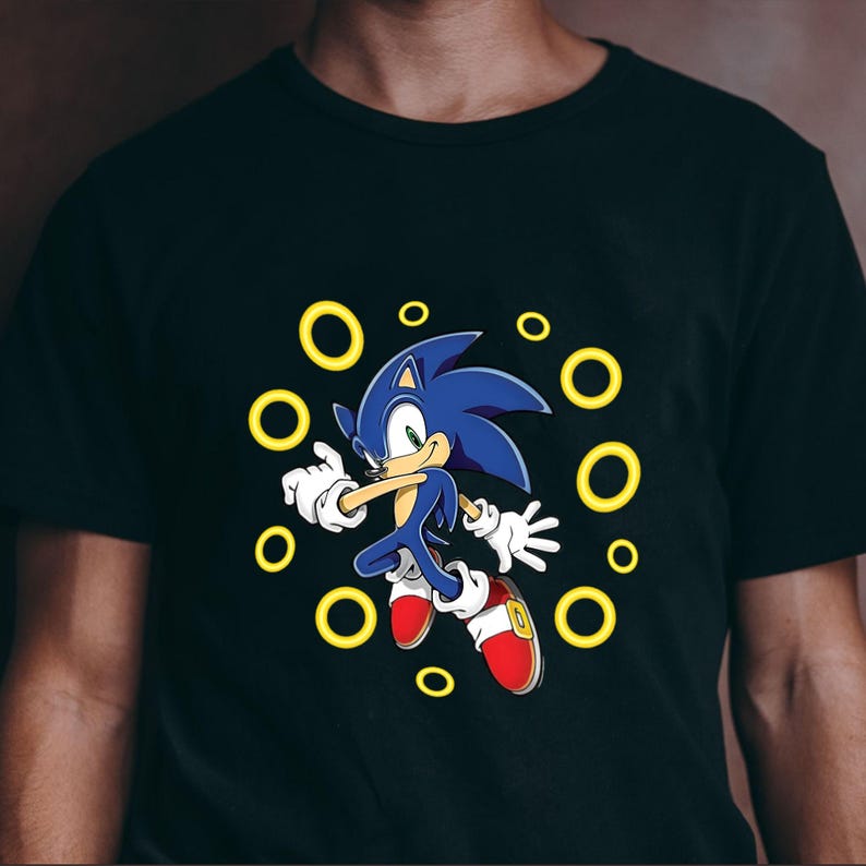 May include: Black t-shirt featuring a cartoon image of Sonic the Hedgehog. The blue hedgehog is surrounded by yellow rings, with red shoes and white gloves. The design is centered on the shirt.