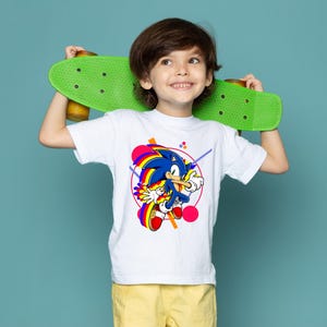 May include: A white t-shirt featuring a colorful graphic of Sonic the Hedgehog. The design includes a blue Sonic character with yellow gloves and red shoes, set against a geometric background. The child is holding a green skateboard.
