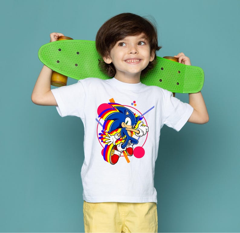 May include: A white t-shirt featuring a colorful graphic of Sonic the Hedgehog. The design includes a blue Sonic character with yellow gloves and red shoes, set against a geometric background. The child is holding a green skateboard.