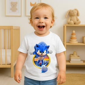 May include: A white t-shirt featuring a vibrant graphic of Sonic the Hedgehog bursting through a golden ring. The blue character is depicted in motion, set against a sunset backdrop. The shirt is worn by a young child.