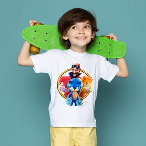 May include: A white t-shirt featuring a colorful graphic of Sonic the Hedgehog and other characters. The child is holding a green skateboard. The background is a solid teal color.