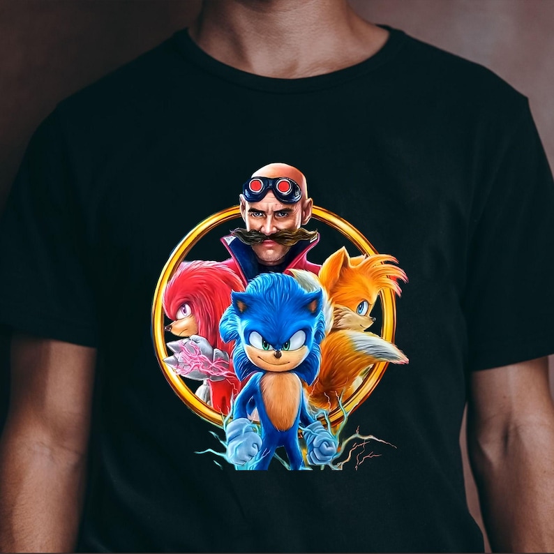 May include: Black t-shirt featuring a colorful graphic of Sonic the Hedgehog and other characters from the video game franchise. The design includes Sonic, Knuckles, Tails, and Dr. Eggman, set within a gold circle.