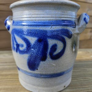 Puede incluir: Un tarro de cerámica blanca con diseños pintados en azul y blanco. El tarro tiene dos asas y un cuerpo redondeado.