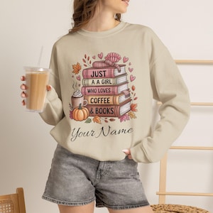 Może przedstawiać: Beżowa bluza z okrągłym dekoltem z grafiką przedstawiającą stos książek, kubek kawy i dynię. Tekst brzmi "JUST A GIRL WHO LOVES COFFEE & BOOKS" oraz "Your Name" napisanym kursywą, w otoczeniu jesiennych liści i serc.