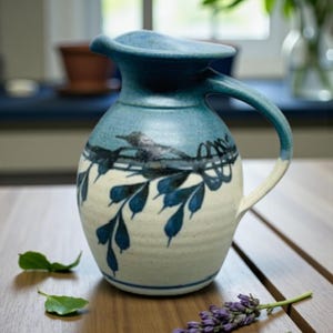 May include: A ceramic pitcher featuring a blue glazed rim and handle that transitions into a cream-colored base. The body is decorated with a dark blue painted design of a bird perched on a branch with leaves, encircling the center of the vessel.