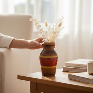 May include: A brown and red ceramic vase with dried flowers, set on a wooden table. The vase has a striped design with a red band in the middle. The hand of a person is holding the vase.