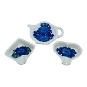 May include: Three white ceramic tea bag holders in different shapes: a teapot, a square, and a heart. Each features a vibrant blue floral design with green accents. The set is ideal for serving tea.