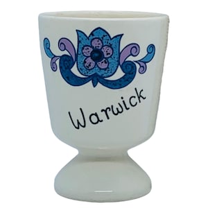 May include: A white ceramic egg cup with a flared base and a floral design in blue and purple. The word "Warwick" is written in black script on the front of the cup.