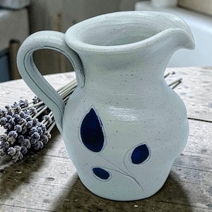 May include: A handmade, off-white ceramic pitcher with a curved handle and a spout. The pitcher features a leaf design with three dark blue teardrop shapes. The pitcher is sitting on a wooden surface.