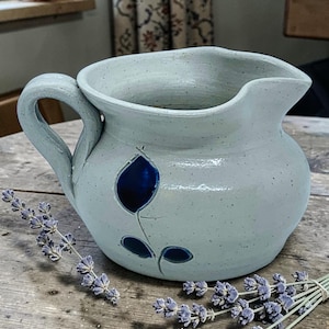 May include: A light blue ceramic pitcher with a handle and spout. The pitcher features a dark blue floral design. A sprig of lavender is placed near the pitcher.