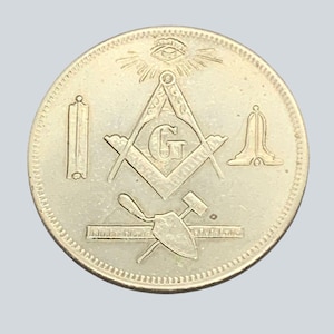 May include: A silver coin featuring Masonic symbols. The coin's design includes a compass, square, and the letter "G" in the center. Other symbols include an all-seeing eye, a trowel, a hammer, and a pillar. The coin has a textured edge.