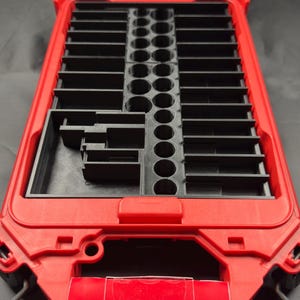 May include: Red and black Milwaukee Packout modular storage organizer. The organizer has a clear lid and a red frame with black interior compartments. The product label reads "MOST VERSATILE DURABLE MODULAR STORAGE SYSTEM."