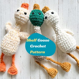May include: Three handmade crochet geese in cream, green, and white, with orange and tan accents. The geese have black eyes and are arranged on a white wooden surface. The text "Shelf Goose Crochet Pattern" is displayed on a blue oval.