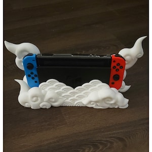 May include: A white 3D printed Nintendo Switch stand shaped like clouds. The stand has a black Nintendo Switch console in it with blue and red joy-cons.