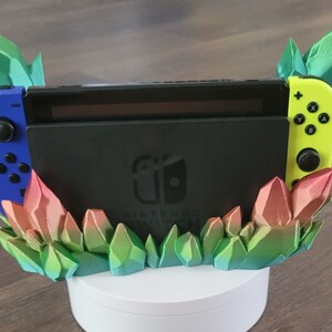 May include: A 3D printed stand for a Nintendo Switch console, featuring a colorful crystal design. The stand is made of multiple layers of green, pink, and blue crystals, creating a vibrant and eye-catching display. The console is placed in the stand, with the controllers on either side.