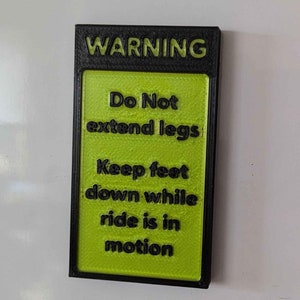 Puede incluir: Un letrero de advertencia negro y verde con el texto "WARNING", "Do Not extend legs", "Keep feet down while ride is in motion".
