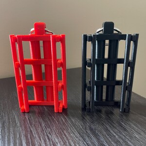 May include: Two 3D-printed keychains. One is bright red, the other is black. Both have a cage-like design with rectangular bars and a ring for keys. They are displayed on a dark wooden surface.