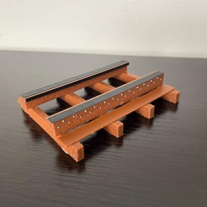 May include: A small, brown wooden bench with a rectangular design. The bench has a series of horizontal slats and vertical supports. The top slats are black with white dots. The bench is designed to hold small items.