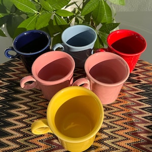 Puede incluir: Seis tazas de cerámica en diferentes colores: azul, azul claro, rojo, rosa, amarillo y azul oscuro. Las tazas están dispuestas en círculo sobre una superficie estampada.