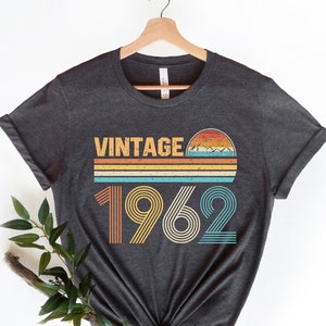 May include: A dark gray t-shirt with a vintage 1962 design. The design features a sunset with mountains and stripes in orange, yellow, blue, and green.