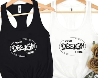 May include: Two racerback tank tops, one black and one white, hanging on wooden hangers. Each tank top features the text "YOUR DESIGN HERE" in a stylized design. The white tank top is adorned with faux flowers and greenery.