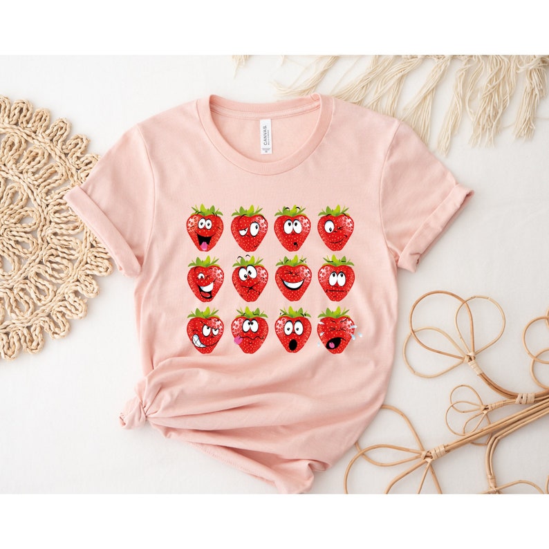 May include: A pink t-shirt with a graphic of 12 strawberries with different facial expressions. The strawberries are red with green leaves.