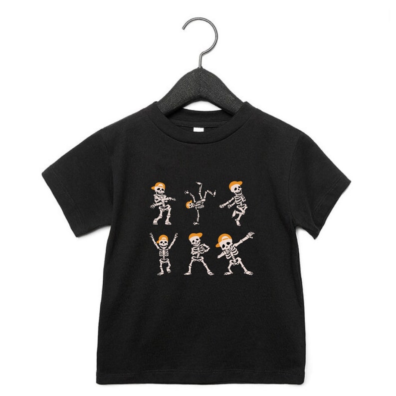 K&ouml;nnte beinhalten: Schwarzes T-Shirt f&uuml;r Kleinkinder mit einem Design von tanzenden Skeletten mit orangefarbenen Kappen. Die Skelette befinden sich in verschiedenen Posen, darunter Breakdance und Dabben. Das T-Shirt h&auml;ngt an einem schwarzen Kleiderb&uuml;gel.