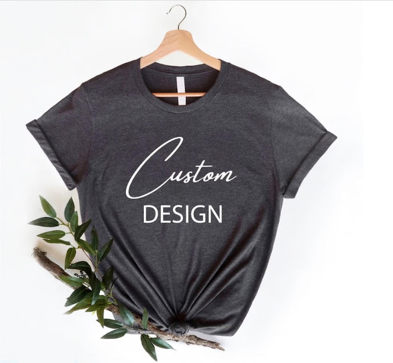 May include: A dark grey t-shirt with a white design that says "Custom DESIGN". The shirt is on a hanger and has a relaxed fit.