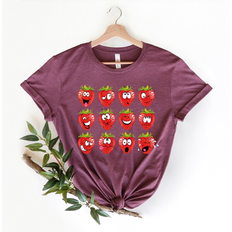 May include: A maroon t-shirt with a graphic of 12 strawberries with different facial expressions. The strawberries are red with green leaves.