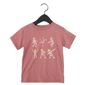 K&ouml;nnte beinhalten: Mauvefarbenes T-Shirt mit einem wei&szlig;en Aufdruck von tanzenden Skeletten mit gelben H&uuml;ten. Die Skelette befinden sich in verschiedenen Posen, darunter Breakdance und Dab. Das Design ist auf einem mauvefarbenen Hintergrund gedruckt.
