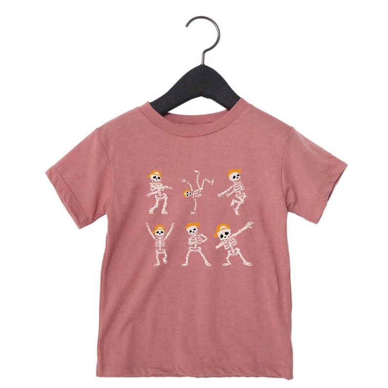 K&ouml;nnte beinhalten: Mauvefarbenes T-Shirt mit einem wei&szlig;en Aufdruck von tanzenden Skeletten mit gelben H&uuml;ten. Die Skelette befinden sich in verschiedenen Posen, darunter Breakdance und Dab. Das Design ist auf einem mauvefarbenen Hintergrund gedruckt.