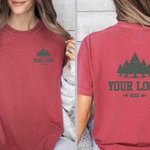 Puede incluir: Camiseta roja de manga corta con un gr&aacute;fico de bosque y el texto "YOUR LOGO HERE" en verde oscuro. El dise&ntilde;o est&aacute; en la parte delantera y trasera de la camiseta. La camiseta es de un material suave.