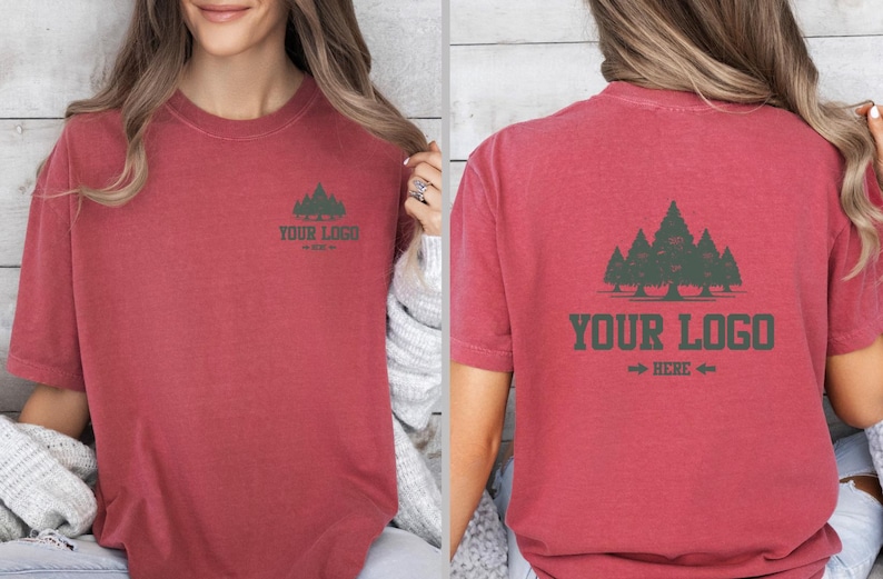 Puede incluir: Camiseta roja de manga corta con un gr&aacute;fico de bosque y el texto "YOUR LOGO HERE" en verde oscuro. El dise&ntilde;o est&aacute; en la parte delantera y trasera de la camiseta. La camiseta es de un material suave.