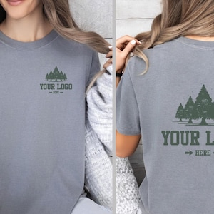 May include: Gray short-sleeved t-shirt with a forest graphic and the text "YOUR LOGO" with an arrow. The shirt is made of a soft material. The design is on the front and back of the shirt.