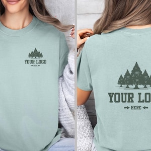 Puede incluir: Camiseta verde claro con un dise&ntilde;o de bosque en la parte delantera y trasera. La parte delantera tiene un peque&ntilde;o dise&ntilde;o de bosque y el texto "YOUR LOGO - HERE -". La parte trasera tiene un dise&ntilde;o de bosque m&aacute;s grande y el texto "YOUR LOGO - HERE -".