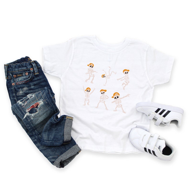 K&ouml;nnte beinhalten: Wei&szlig;es T-Shirt mit Skelett-Design, kombiniert mit blauen Jeans und wei&szlig;en Turnschuhen. Das T-Shirt zeigt mehrere Cartoon-Skelette mit orangefarbenen Haaren und H&uuml;ten in verschiedenen Posen. Die Jeans sind ausgewaschen und die Turnschuhe haben schwarze Streifen.