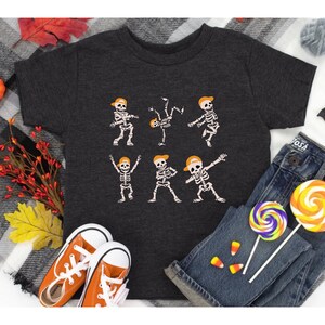 K&ouml;nnte beinhalten: Dunkelgraues T-Shirt mit einem Skelett-Design mit orangefarbenen Kappen in verschiedenen Tanzposen. Das Shirt wird mit orangefarbenen Turnschuhen, blauen Jeans und bunten Lollis kombiniert, was ein verspieltes Halloween-Thema ergibt.