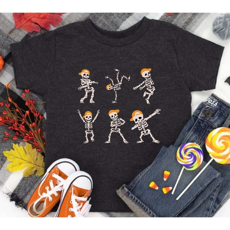 K&ouml;nnte beinhalten: Dunkelgraues T-Shirt mit einem Skelett-Design mit orangefarbenen Kappen in verschiedenen Tanzposen. Das Shirt wird mit orangefarbenen Turnschuhen, blauen Jeans und bunten Lollis kombiniert, was ein verspieltes Halloween-Thema ergibt.