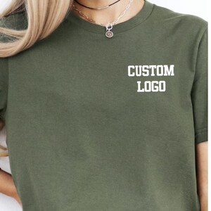 May include: Olive green t-shirt with the text "CUSTOM LOGO" printed in white. The shirt is a crew neck style and appears to be made of a soft material. The shirt is a solid color and has short sleeves.