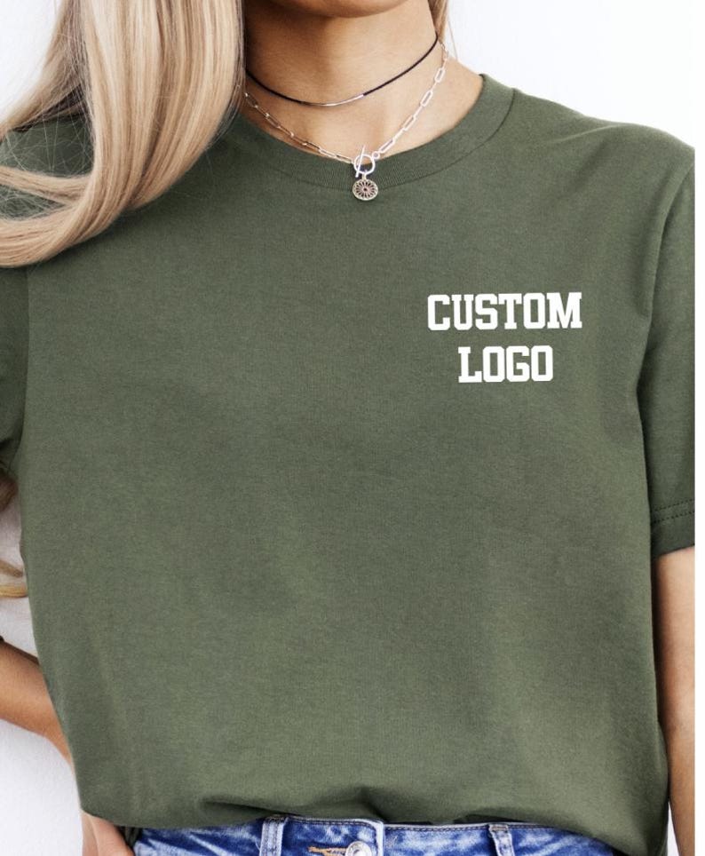 May include: Olive green t-shirt with the text "CUSTOM LOGO" printed in white. The shirt is a crew neck style and appears to be made of a soft material. The shirt is a solid color and has short sleeves.
